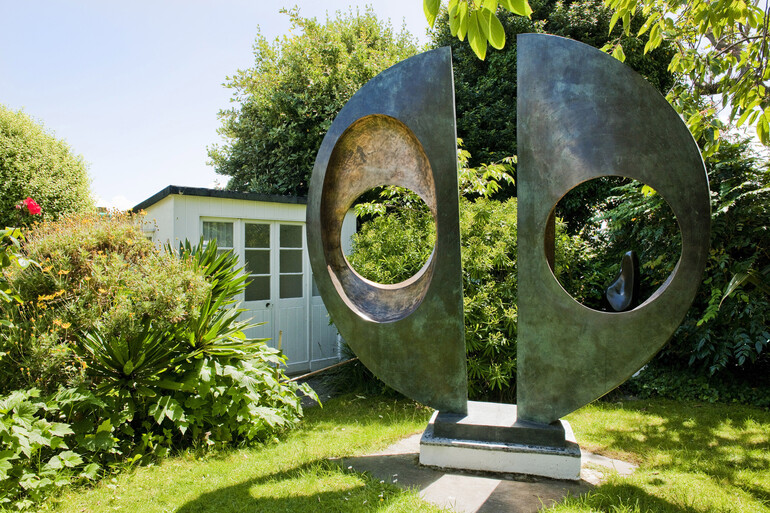 A large Barbra Hepworth sculpture of two semi circles stands outside a white shed next to hedges