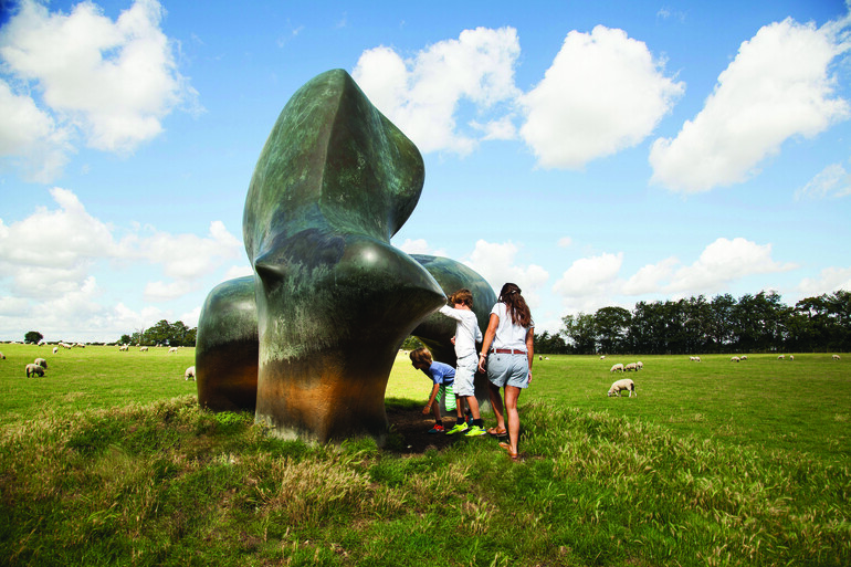 Children explore inside a large Henry Moore sculpture standing on a field with sheep grazing in the background