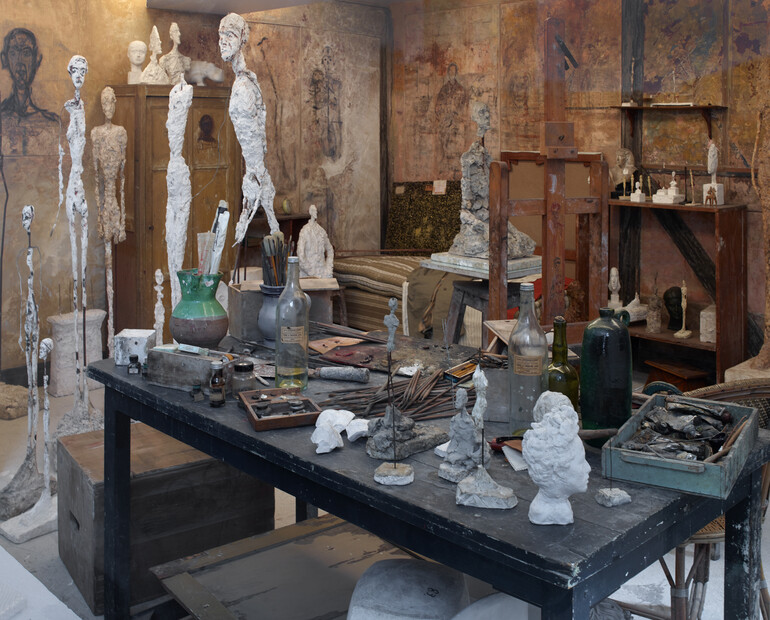A messy sculpture studio, in the foreground is a cluttered bench and surrounding this are half finished models of Giacometti's works