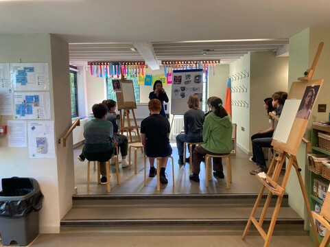 Sarah Thien stands in front of a group of young people sat on stools in the Clore learning studio. The white board next to her contains various pictures of portraiture