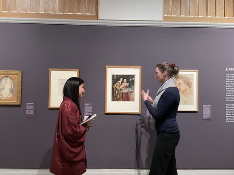 Two women stand in front of a painting, one, in a blue jumper raises her hands as she talks while the other, in a red blouse, listens attentively