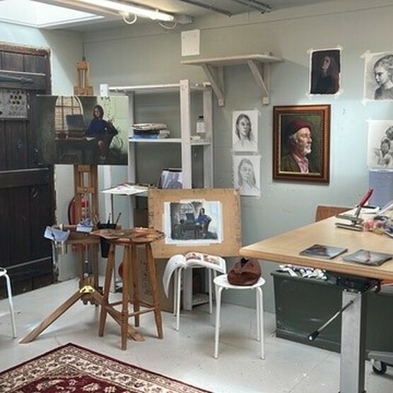 An artist's studio containing an easel and several stools, and a table covered in art supplies. On the wall are several portraits and pictures of G F Watts work