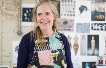 Mary smiles to the camera, in the background are artworks pinned on the wall