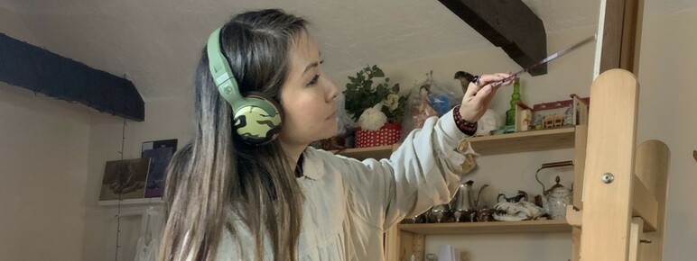 Sarah wears headphones and works on a painting on an easel