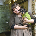 Ashleigh smiles at the camera and holds small black and brown dog, Merlin. Merlin wears a bright green jumper.
