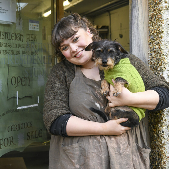 Ashleigh smiles at the camera and holds small black and brown dog, Merlin. Merlin wears a bright green jumper.