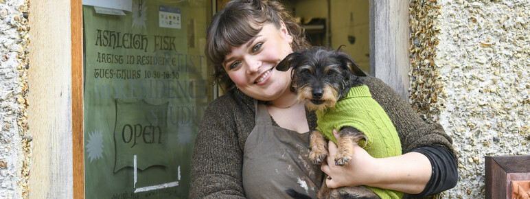 Ashleigh smiles at the camera and holds small black and brown dog, Merlin. Merlin wears a bright green jumper.