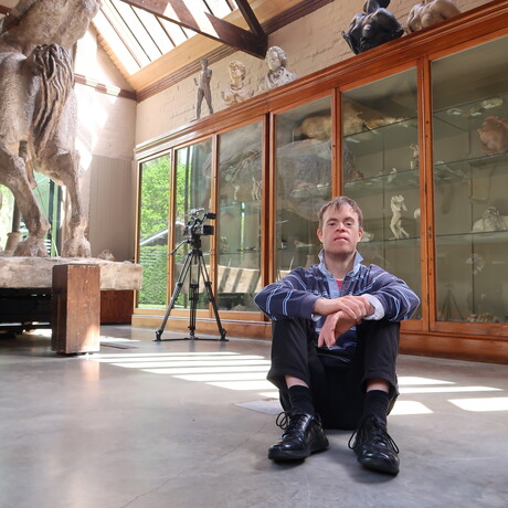Chris sits on the concrete floor in the Sculpture Gallery, the large sculpture Physical Energy and a camera can be seen behind him