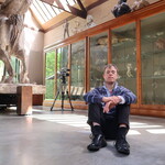 Chris sits on the concrete floor in the Sculpture Gallery, the large sculpture Physical Energy and a camera can be seen behind him