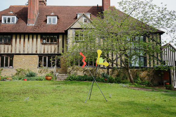 Photo of Limnerslease with an abstract sculpture on the lawn in front. The sculpture consists of red, yellow and green perspex in abstract shapes on metal poles