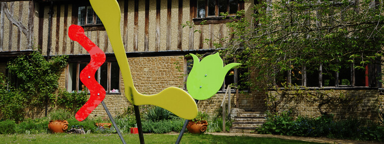 Photo of Limnerslease with an abstract sculpture on the lawn in front. The sculpture consists of red, yellow and green perspex in abstract shapes on metal poles