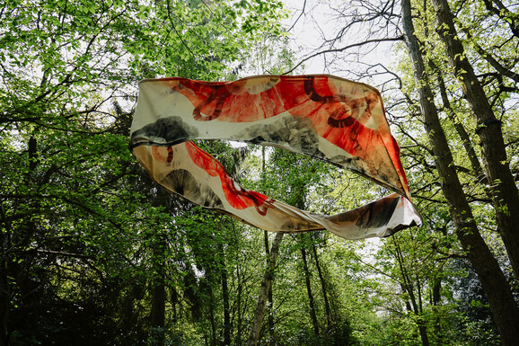 Photo of an abstract sculpture suspended in between trees in a woodland, made from canvas, featuring expressive orange paint marks