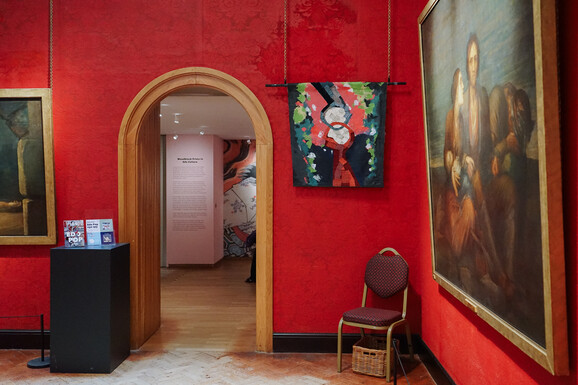 Photo inside the Historic Galleries at Watts Gallery. There is an artwork hung on the wall which incorporates textiles and painting in a collage-style. It is displayed beside paintings by G F Watts