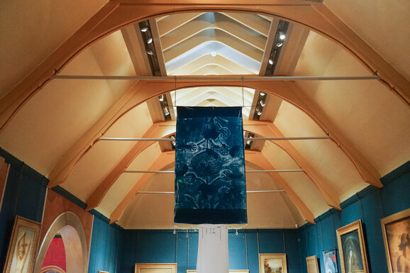 Cyanotype hanging from the ceiling of Watts Gallery with beading decorating the bottom of the fabric. There is an abstract design on the fabric