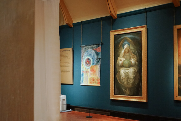 Photo inside the Historic Galleries at Watts Gallery. There is an abstract colourful artwork hung on the wall made from canvas. It is displayed beside paintings by G F Watts