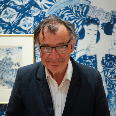 Photo of Frank Milner; a man wearing a suit and glasses with grey hair. He is standing in front of a blue and white backdrop