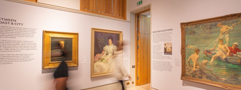 White gallery space with large gold-framed paintings on the wall, two visitors walk past