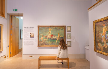 A visitor sits on a bench in front of a large beach scene painting