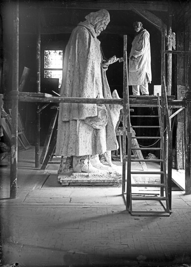 Black and white photo of artist G F Watts on scaffolding next to large statue of Lord Tennyson