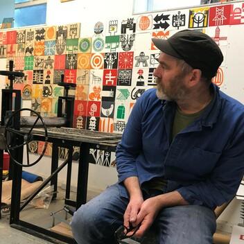 Photograph of a man sitting on a stool in his art studio. There are various prints hung on the wall behind him.