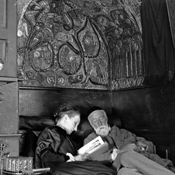 G F and Mary Watts sitting together in the reading nook at Limnerslease. Mary is reading aloud