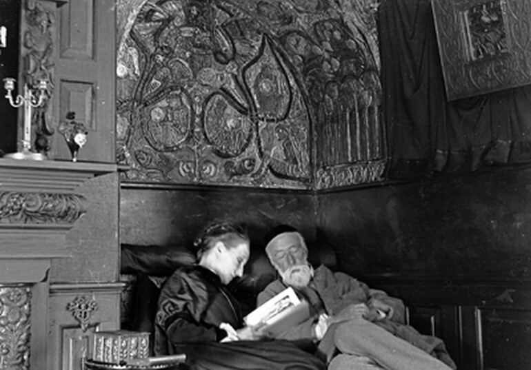 G F and Mary Watts sitting together in the reading nook at Limnerslease. Mary is reading aloud