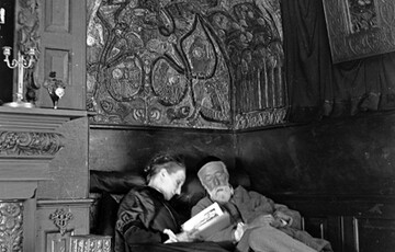 G F and Mary Watts sitting together in the reading nook at Limnerslease. Mary is reading aloud