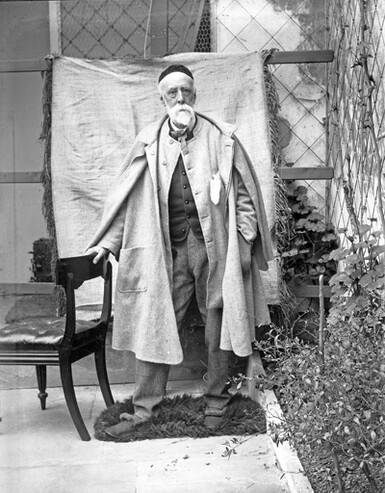 G F Watts posing for a photo portrait. He is dressed in a suit and a large coat.