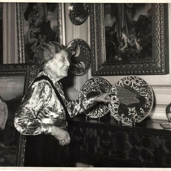 Black and white photo of a woman gesturing to a plate by William De Morgan placed on a mantelpiece