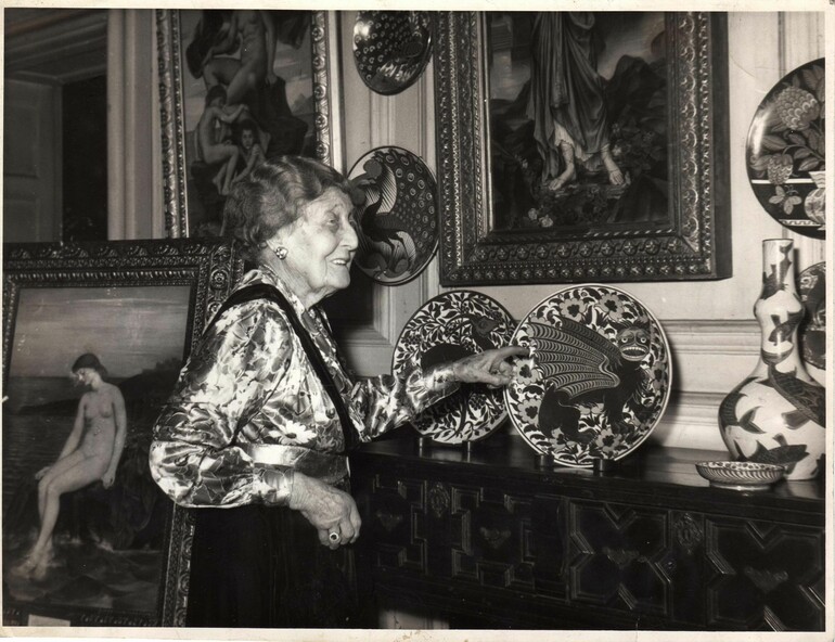 Black and white photo of a woman gesturing to a plate by William De Morgan placed on a mantelpiece
