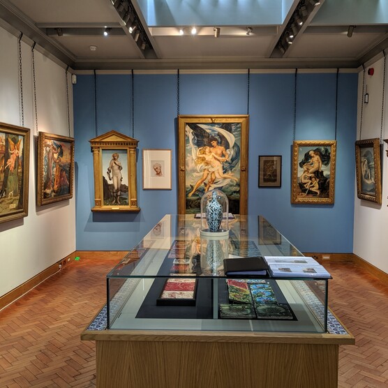A view of the De Morgan Gallery looking towards Evelyn De Morgan's paintings, with a glass display case containing works by William De Morgan in the centre of the room