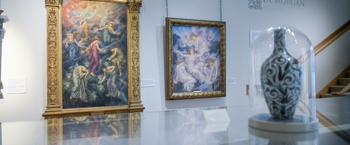 A floral vase encased in a glass display case with two colourful oil paintings in gold frames in the background