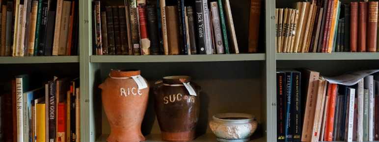 An image of Watts Gallery's archive room. There is sage green selving units full of different coloured books. In shelving at the bottom is two large ceramic pots. One orange rice pot and one brown sugar pot.