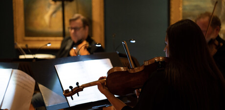 A photograph of a woman playing the violin looking at sheet music with two men in the background also sitting playing the violin