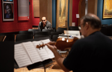 A photograph of composer, Des Oliver looking at a man playing the violin whilst reading sheet music