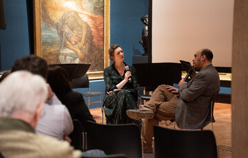 A photograph of curator Emily Burns and composer Des Oliver sitting in the Historic Galleries holding microphones talking