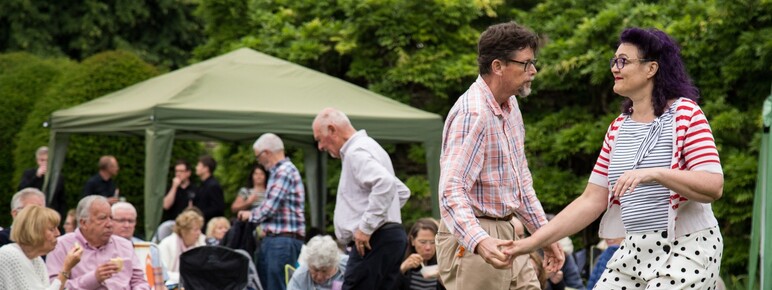 A photograph of a man and a women outside dancing on grass with an audience watching them