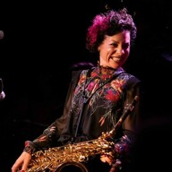 A photograph of a women smiling with brown and pink hair wearing a black dress holding a saxophone
