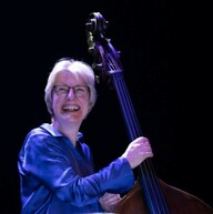A photograph of a women smiling wearing glasses and a blue shirt looking at the camera holding a cello