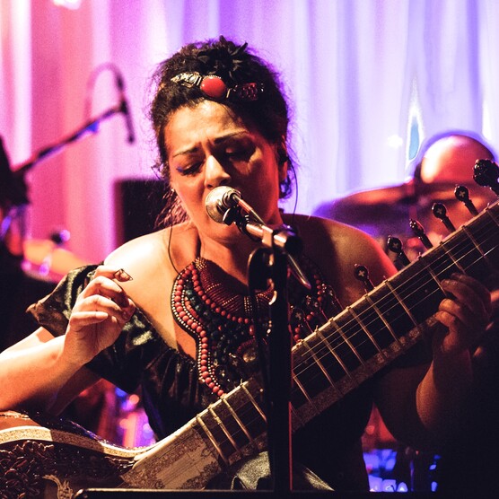 A woman playing the sitar singing into a mic. Behind her is a pink curtain and pink and purple lighting
