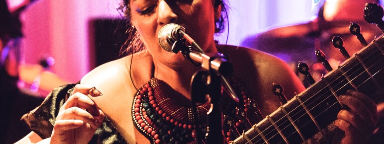 A woman playing the sitar singing into a mic. Behind her is a pink curtain and pink and purple lighting