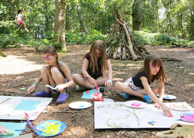 A photograph of three girls sitting on the ground in the woods at Watts Gallery colouring and painting on paper