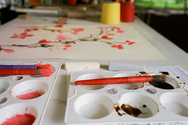 Photo of paintbrushes in a paint palette with a cherry blossom painting placed behind them