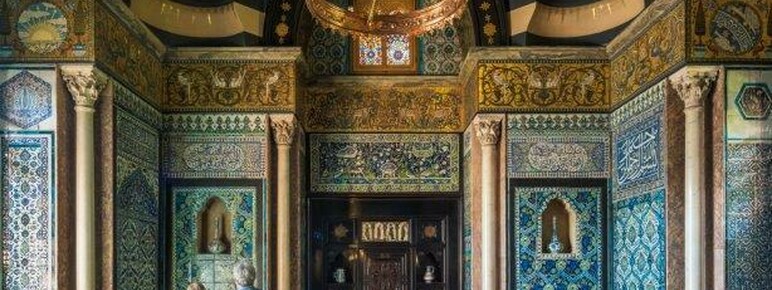 An ornate tiled hall with a small pool in the centre surrounded by rugs. Two people stand looking upwards to the left