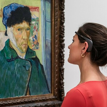 A photograph of a women wearing a headset looking at a Van Gogh painting hung on the wall