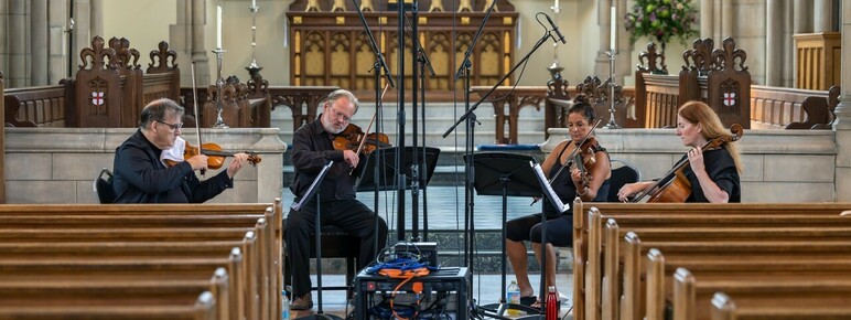 Two men and a woman playing violin's whilst reading music and another woman playing the cello whilst sat inside a church