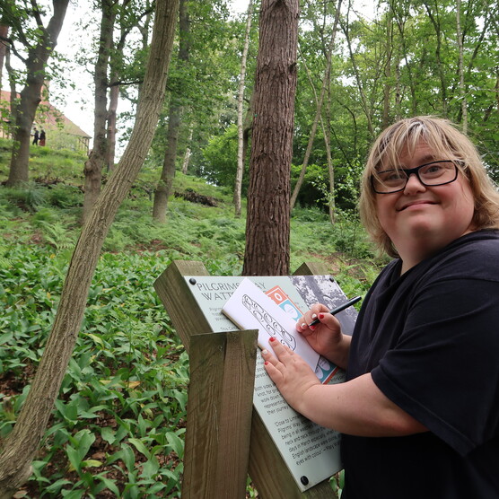 A woman draws while leaning on a information plaque in Limnerslease woodland, she smiles towards the camera