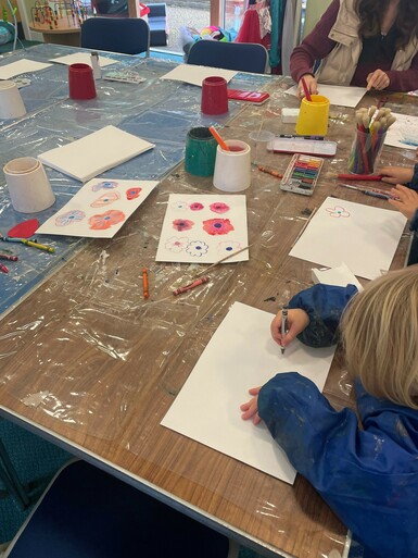 A child in a blue smock draws flowers at a table covered in other drawings and art supplies like watercolour sets