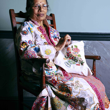 An elderly woman wearing an embroidered gown, sat in a high-backed wooden chair