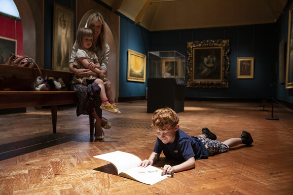 A boy lies on the gallery floor reading while his mother sits on a bench watching him, holding another young child.